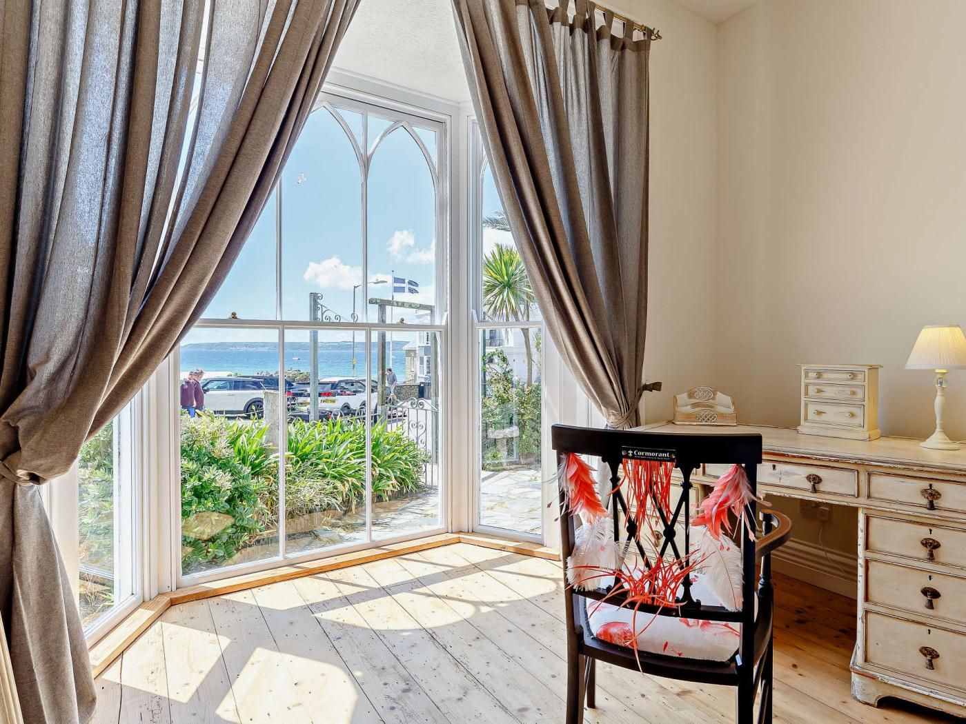 Ein Zimmer mit Blick auf eine Küstenlandschaft, einem Klavier, einem Schreibtisch mit Schubladen und einem Sessel mit Blumenmuster.