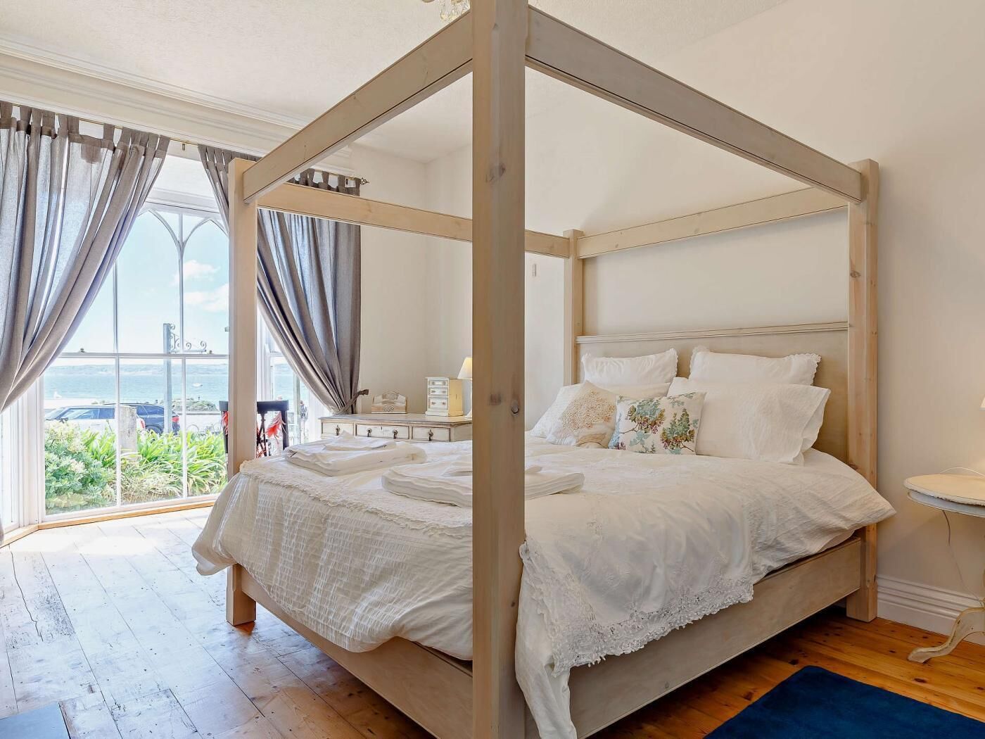 Un lit à baldaquin avec une structure en bois, une literie blanche et une vue sur l’extérieur grâce à de grandes fenêtres.