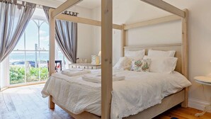 Una cama con dosel de madera, ropa de cama blanca y vistas al exterior a través de amplios ventanales.