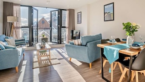 Un salon moderne avec un canapé, un fauteuil et une table à manger pour deux personnes, de grandes fenêtres offrant une vue sur la ville, et un téléviseur à écran plat posé sur un meuble.