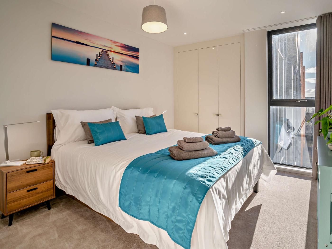 A bedroom with a large bed, a nightstand, a bedside table, a window with a view, and a painting on the wall.