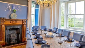 Een eetkamer met een lange tafel gedekt voor een maaltijd, een open haard en grote ramen.