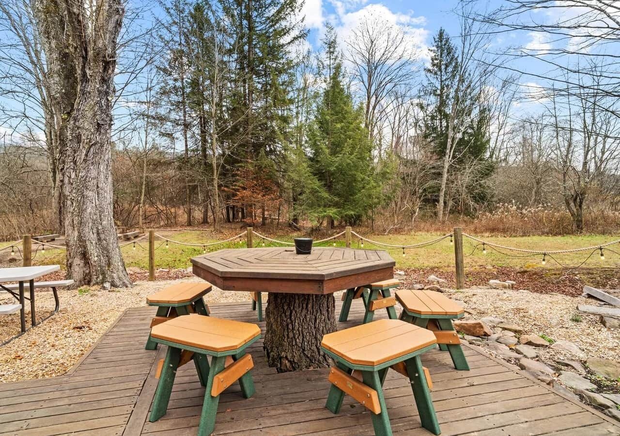 A circular wooden table with benches, surrounded by a wooden deck, set in a wooded area.