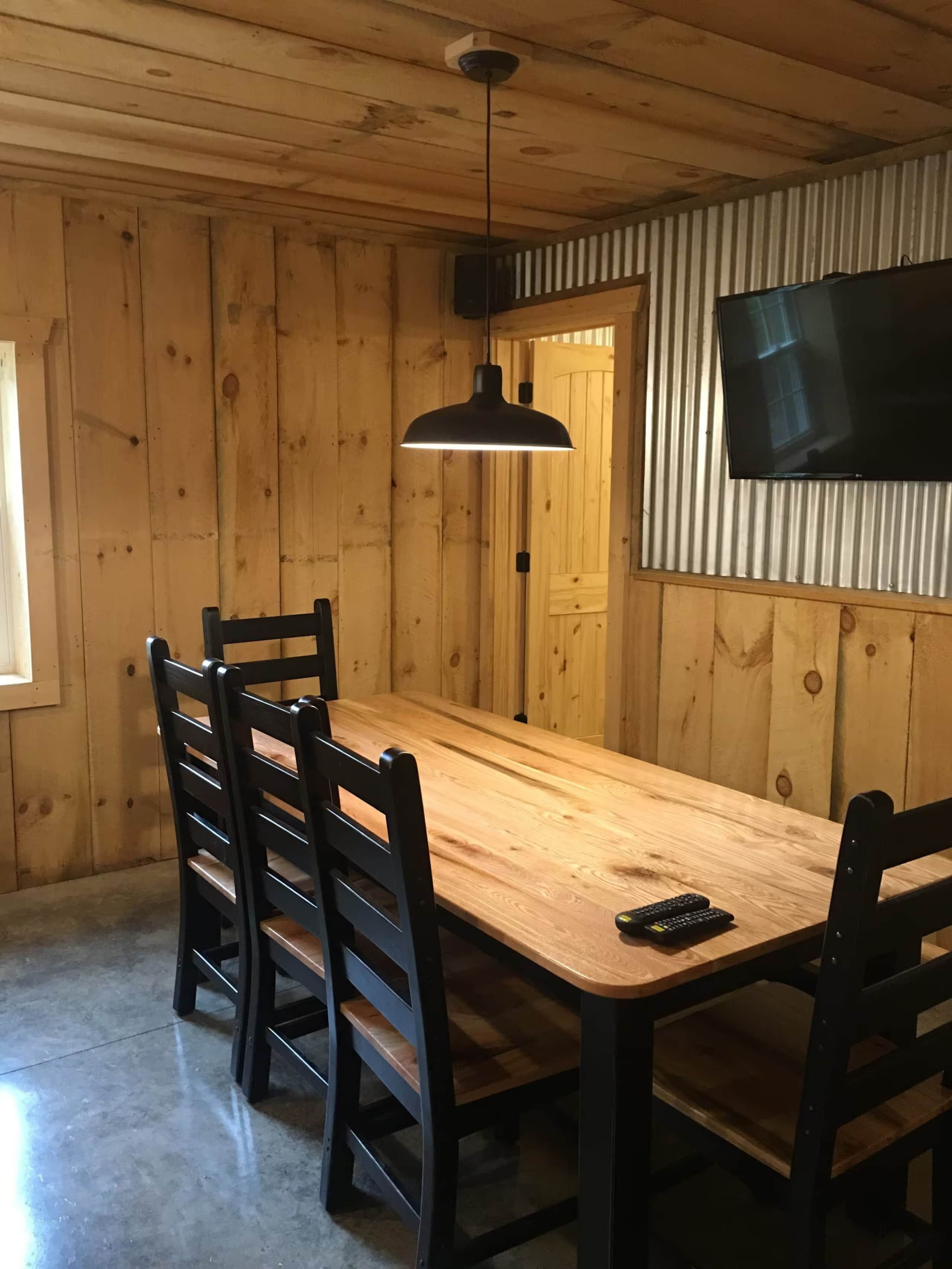 Una habitación con paneles de madera, una mesa larga, cuatro sillas, una lámpara en el techo y un televisor montado en la pared.
