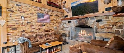 A cozy living room with a stone fireplace, a wooden ceiling, and a brick wall.