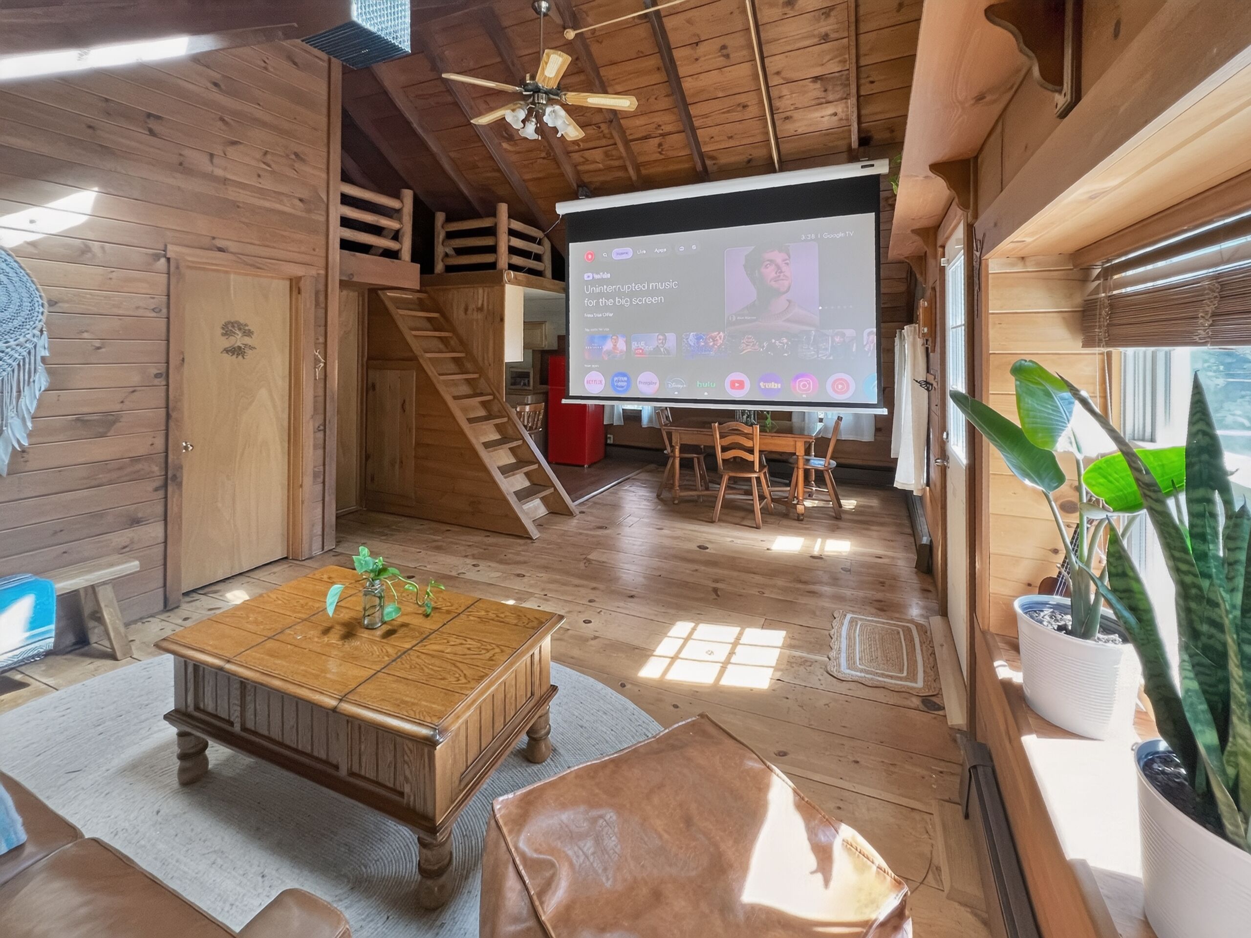 A wooden interior with a large screen displaying a music video, a staircase, and a dining area.
