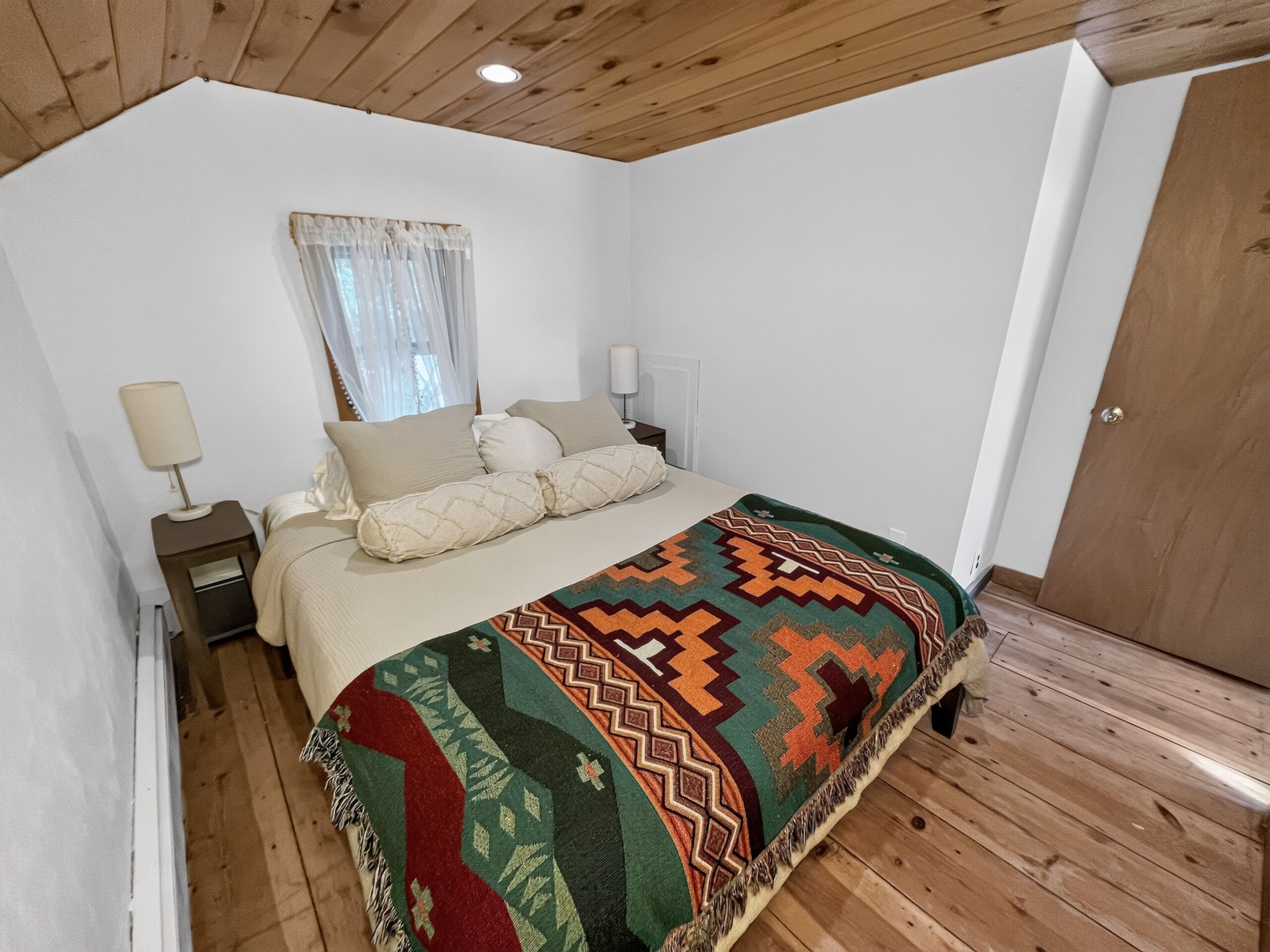 A bedroom with a wooden ceiling, a bed with a patterned blanket, a nightstand with a lamp, and a window with curtains.