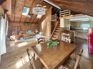 Interior de uma cabana de madeira com sala de jantar, cozinha e escada para um andar superior.