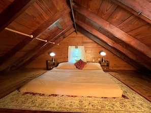 Quarto no sótão com uma cama grande, duas luminárias de cabeceira e uma janela com cortinas.