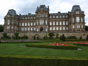 A large, historic building with symmetrical architecture, multiple windows, and a formal garden with hedges and flower beds.