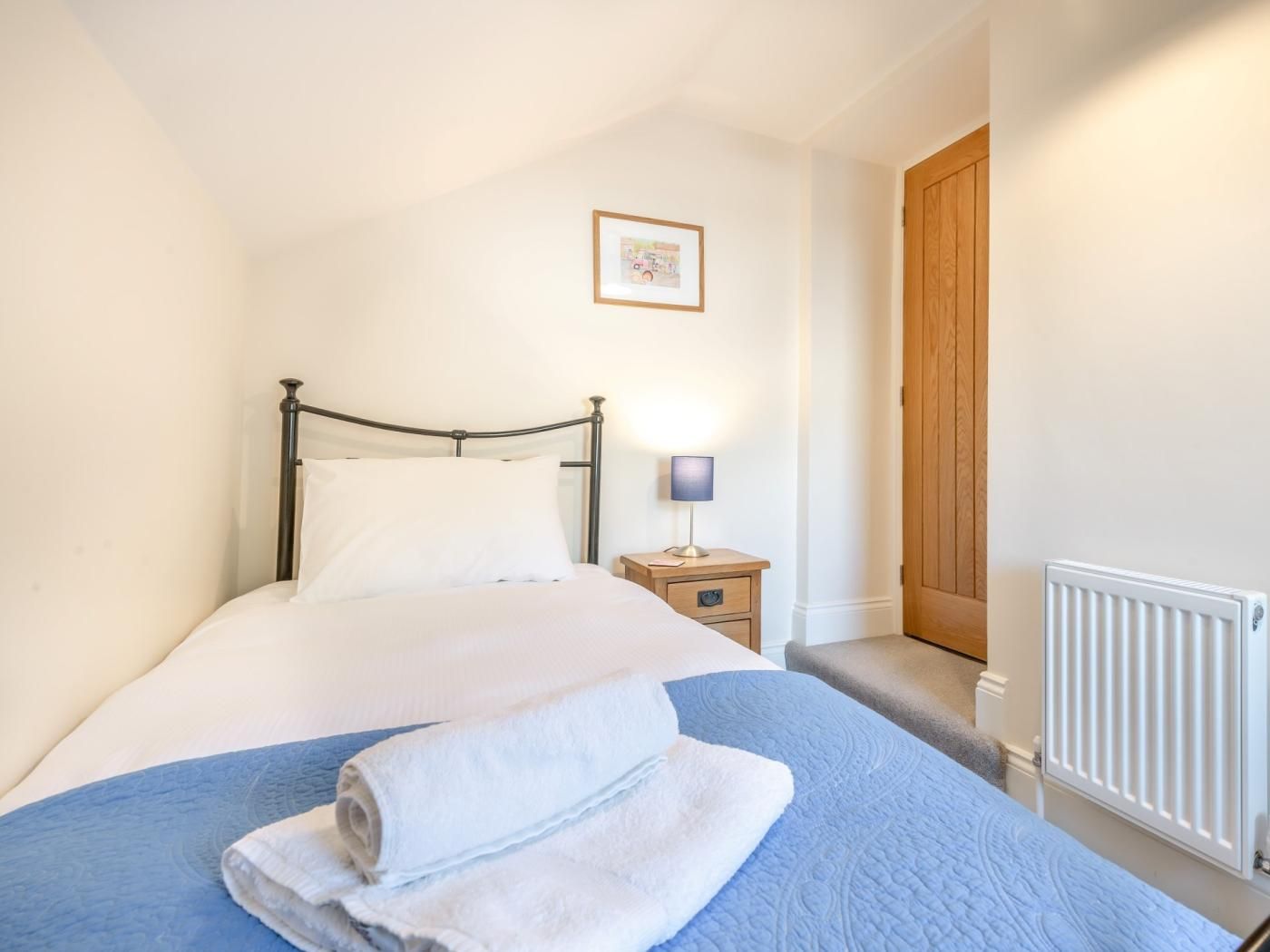 A single bed with a blue quilt, a white pillow, and a rolled white towel. There is a bedside table with a lamp, a framed picture on the wall, and a wooden door.