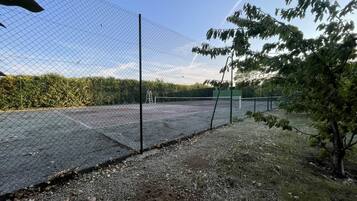 Un court de tennis entouré d’une clôture et doté d’un filet, bordé d’arbres et d’une haie.