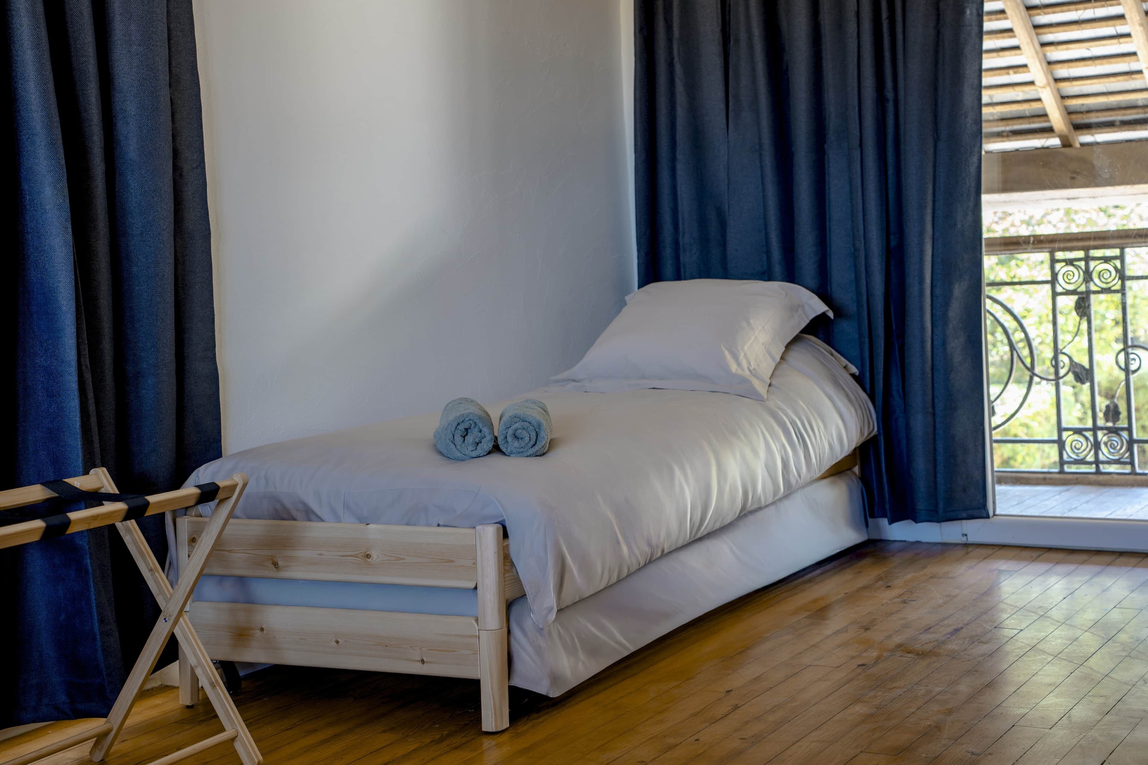 Una cama individual con estructura de madera, ropa de cama blanca y dos toallas enrolladas.