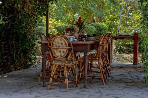 A wooden dining table with chairs, surrounded by greenery and a brick structure.