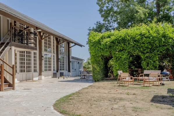 Un bâtiment doté de grandes fenêtres et d’un espace extérieur couvert, aménagé avec des chaises et un barbecue.