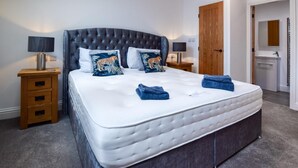 Ein Schlafzimmer mit einem großen Bett, zwei Nachttischen mit Lampen, einer Holztür und einem Badezimmer mit Waschbecken und Spiegel.