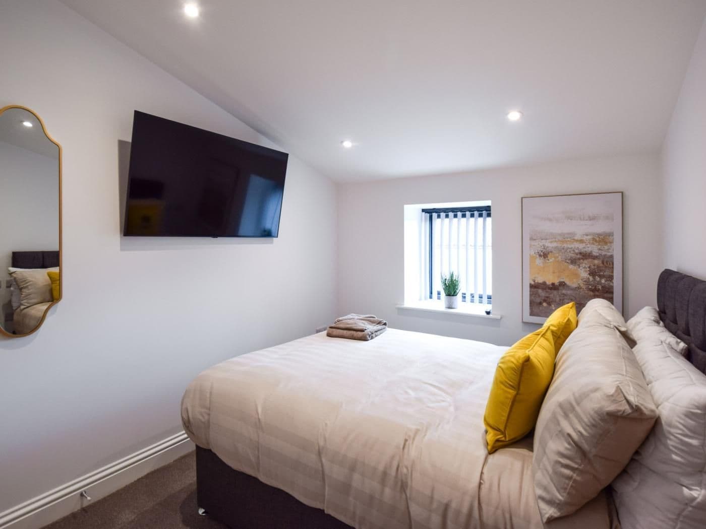 A bedroom with a bed, a flat-screen TV mounted on the wall, a window with blinds, and a framed picture on the wall.