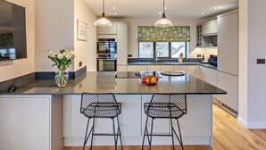 A modern kitchen with a central island, two bar stools, pendant lighting, and a large window. - 1 Bed in Woolacombe (oc-93920) (Woolacombe)
