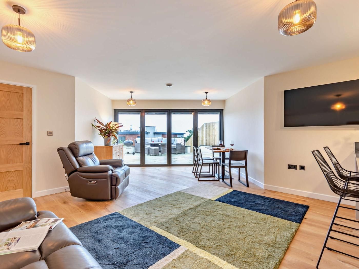 Een moderne woonkamer met een leren bank, een eethoek met een tafel en stoelen, een groot raam en een flatscreen televisie aan de muur.