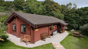Eine kleine, eingeschossige Holzhütte mit geneigtem Dach, einem Schotterweg und einem Picknicktisch.