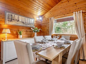 A dining area with a wooden table set for four, white chairs, a vase with flowers, and a painting on the wall. - 3 Bed in Dalbeattie (oc-93985) (Sandyhills)