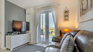 Ett vardagsrum med en soffa i läder, en platt-TV, ett vitt skåp och en dörr som leder ut till ett utomhusområde.