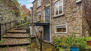 A stone building with a staircase leading to a door, surrounded by greenery and a stone pathway.
