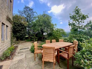 A stone patio with a wooden table and chairs, surrounded by lush greenery and trees. - 6 Bed in Huddersfield (oc-93984) (Huddersfield)