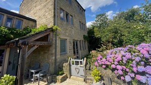 A stone building with a covered patio, outdoor seating, and a garden with hydrangeas. - 6 Bed in Huddersfield (oc-93984) (Huddersfield)