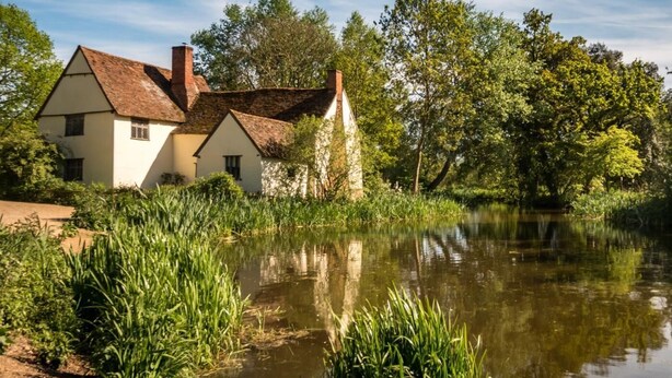 A traditional house with a white facade and a red-tiled roof, situated beside a calm body of water with lush greenery.