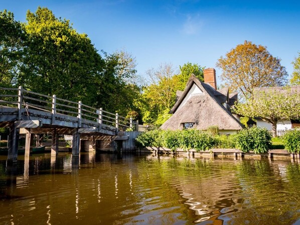 A thatched-roof house by a calm river with a wooden bridge.