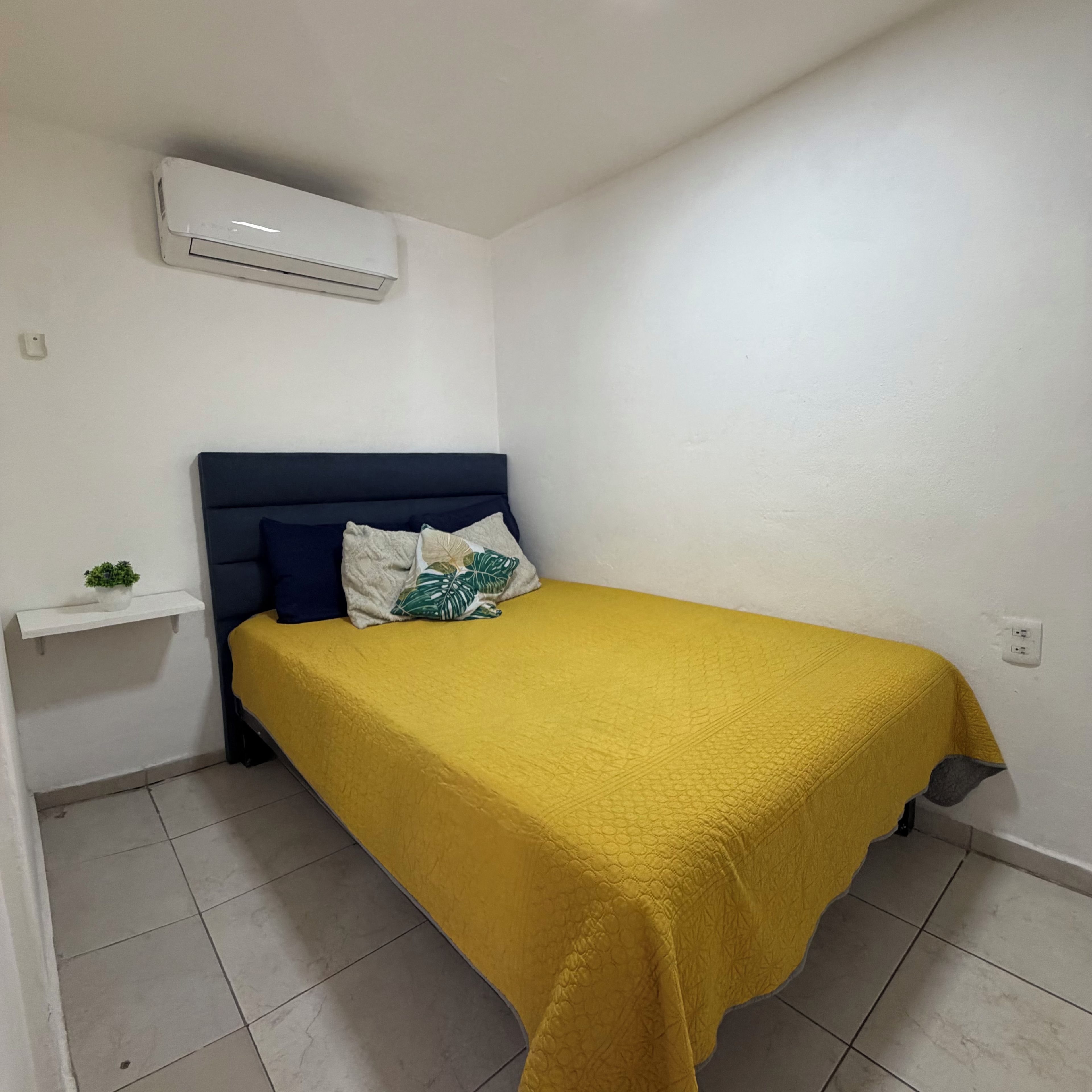 Una cama individual con una colcha amarilla, cabecera oscura y dos almohadas. Se observa un equipo de aire acondicionado blanco encima de la cama.