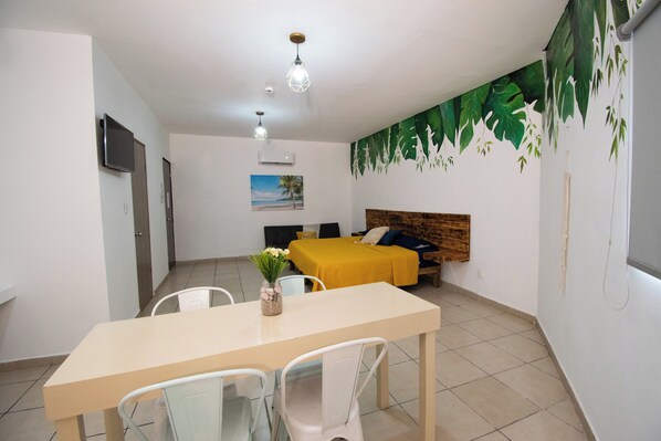 Una habitación con cama, una mesa y sillas, televisor y un mural tropical en la pared.