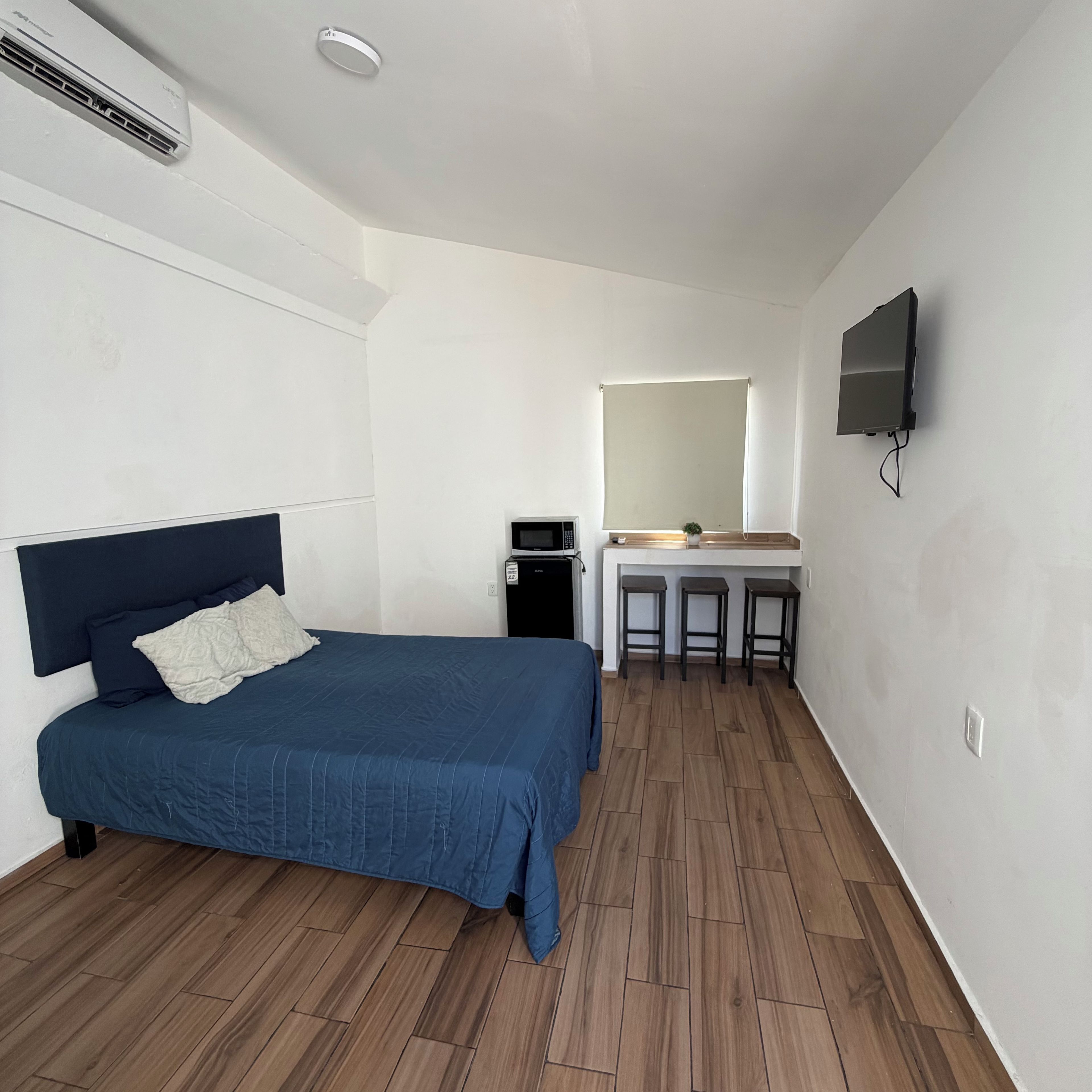 A bedroom with a bed, a desk, a chair, a television, and a small kitchenette.