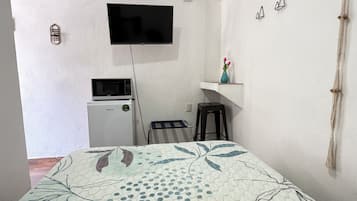 Habitación con una cama, televisión montada en la pared, microondas, un taburete pequeño y un estante con un florero.