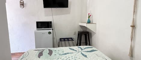 A room with a bed, a TV mounted on the wall, a microwave, a small stool, and a shelf with a vase.