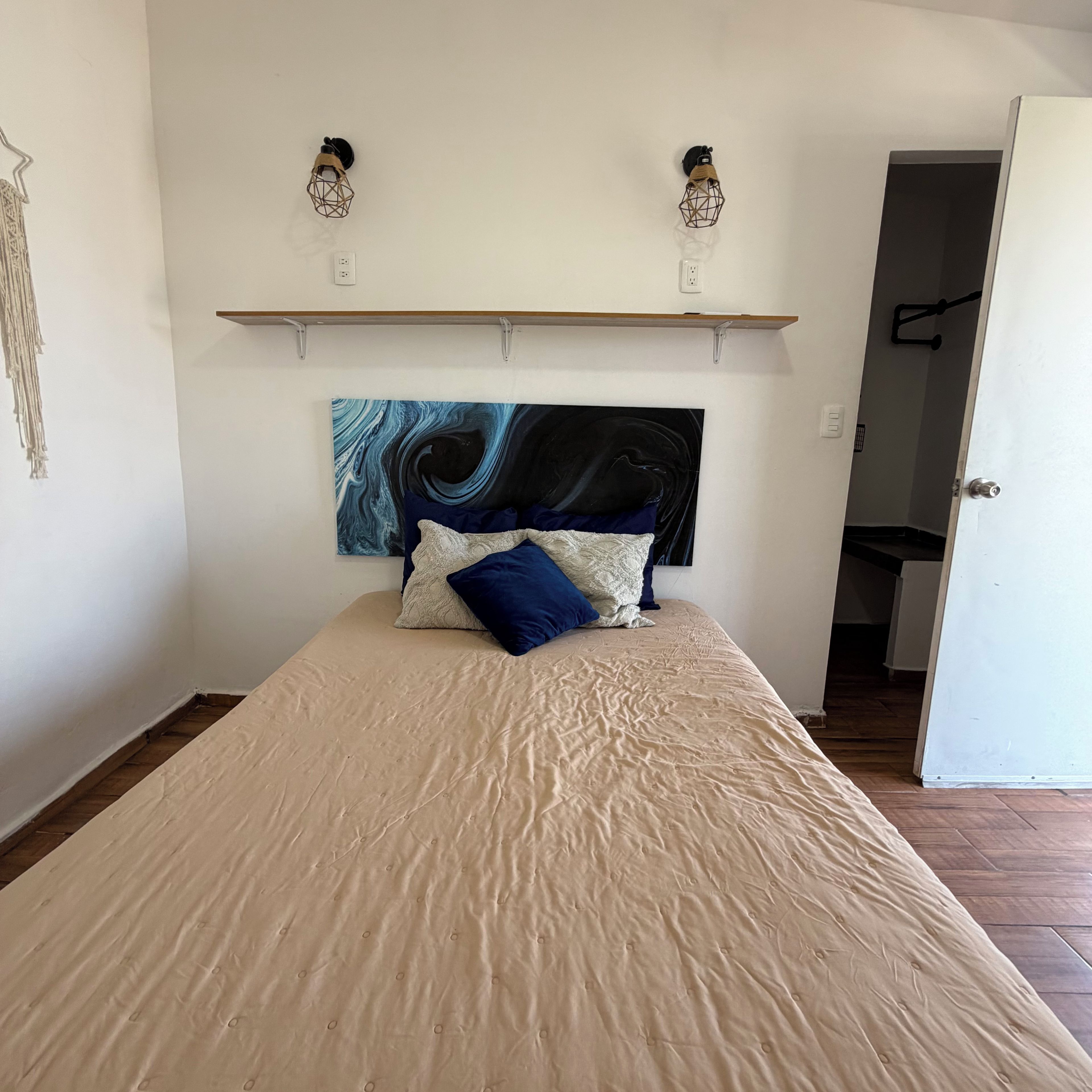 Un dormitorio con cama, cabecera con un cuadro y dos lámparas fijadas a la pared.