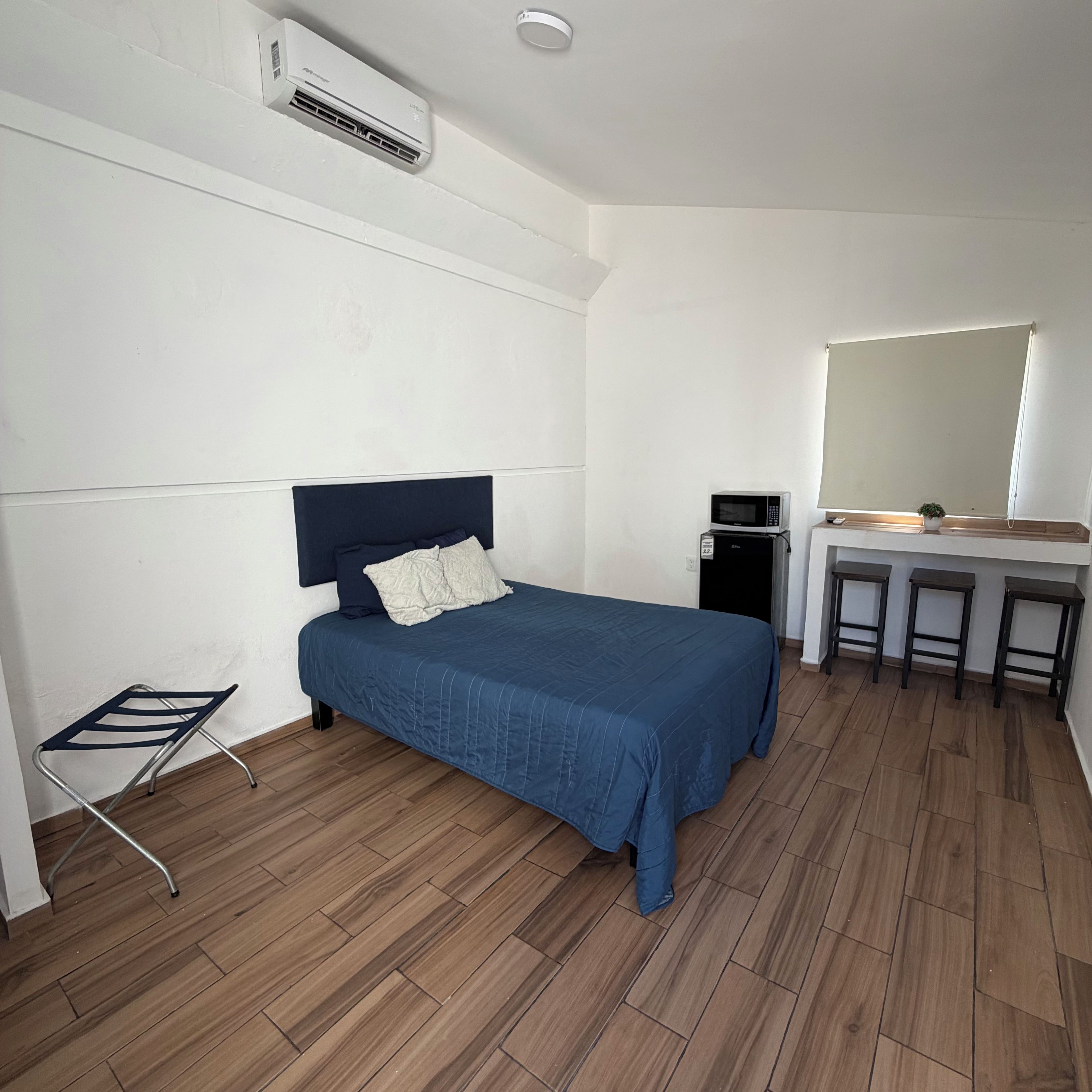 Una habitación con una cama, un escritorio con espejo, un taburete pequeño, un microondas y un equipo de aire acondicionado.