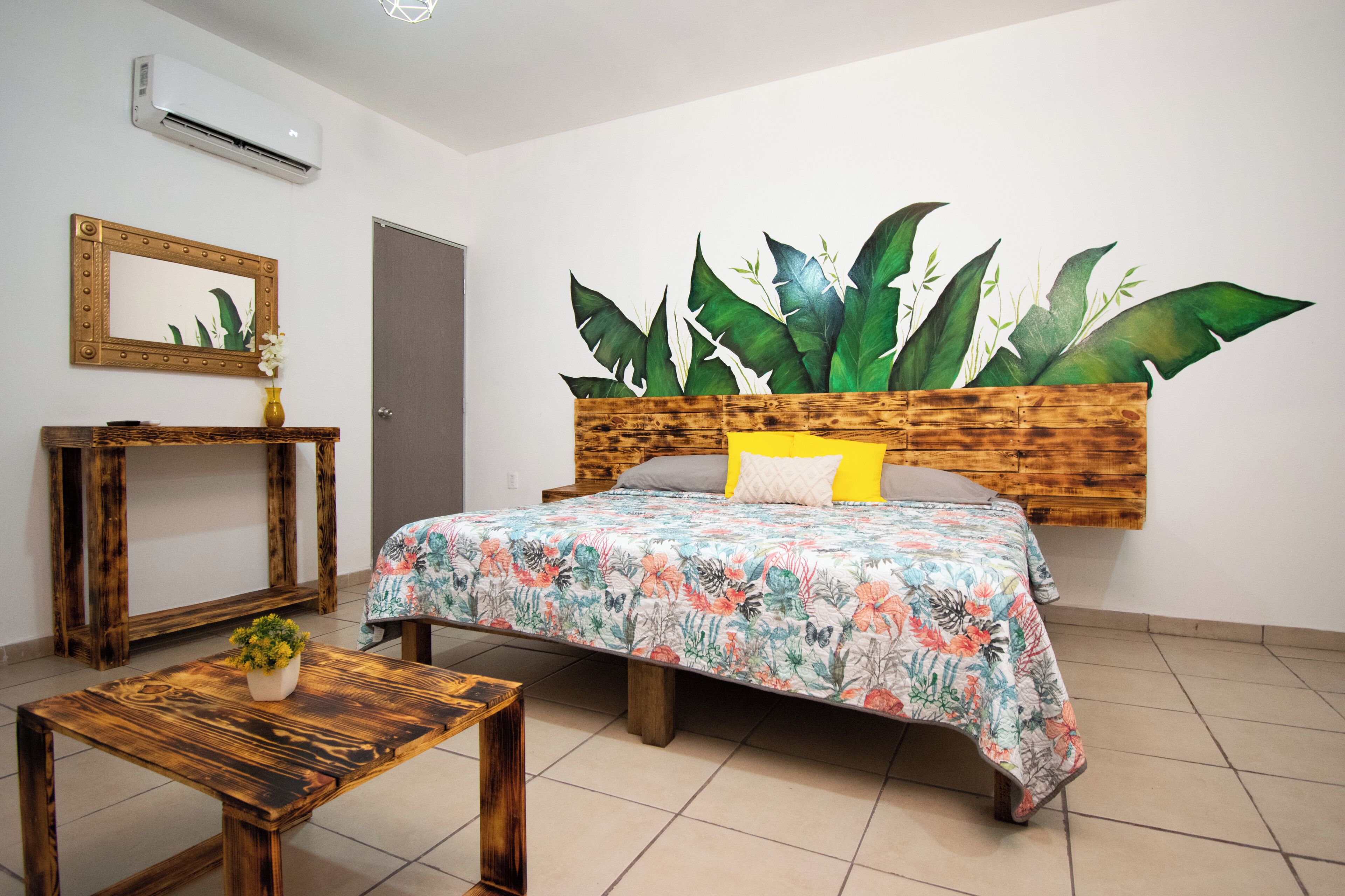 A bedroom with a bed, a wooden headboard, a bedside table, a small potted plant, and a wall-mounted air conditioner.