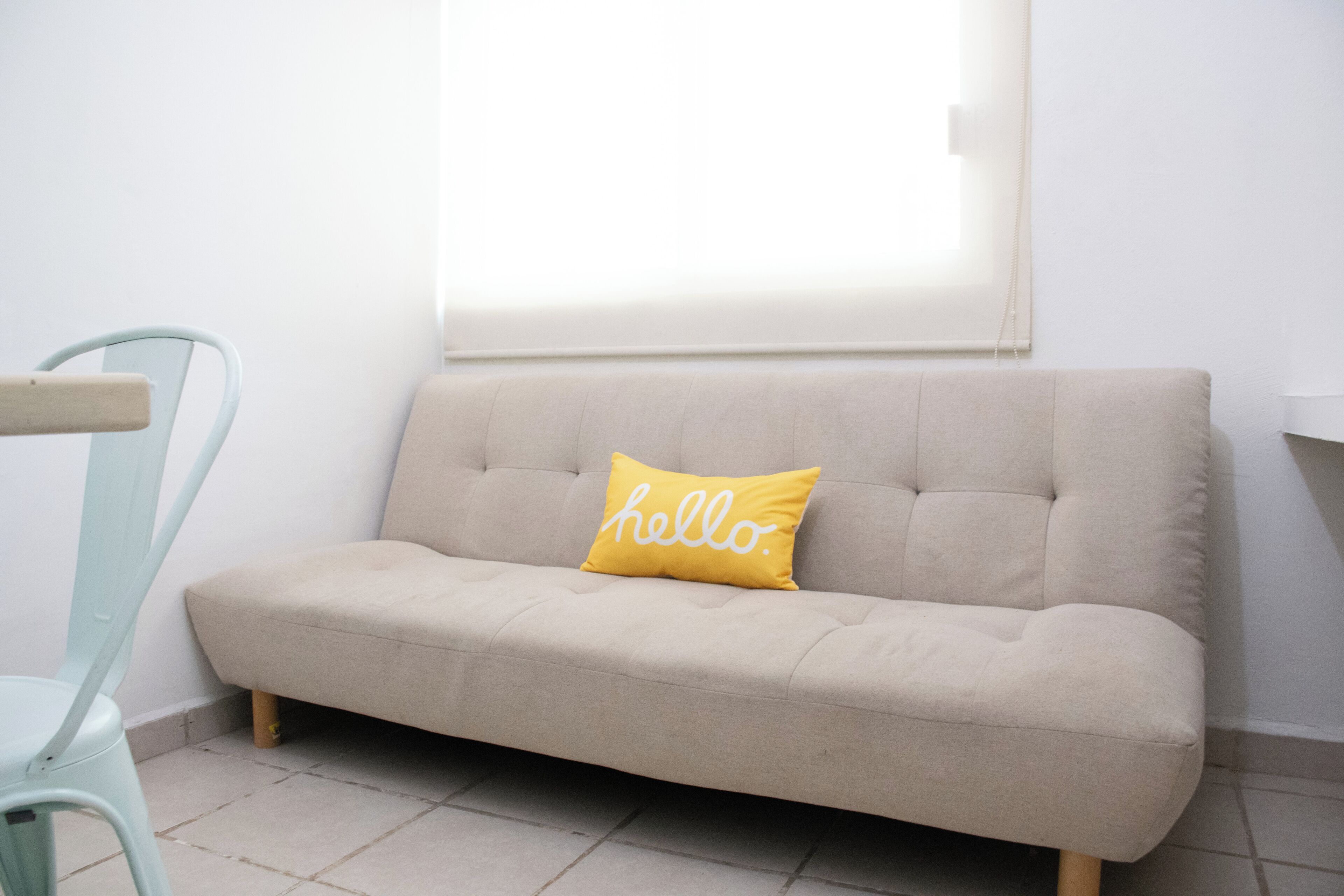 Un sofá de tonos claros con un cojín que tiene la palabra 'hello' escrita, ubicado frente a una pared con una ventana arriba.