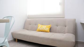 Un sofá de tonos claros con un cojín que tiene la palabra 'hello' escrita, ubicado frente a una pared con una ventana arriba.
