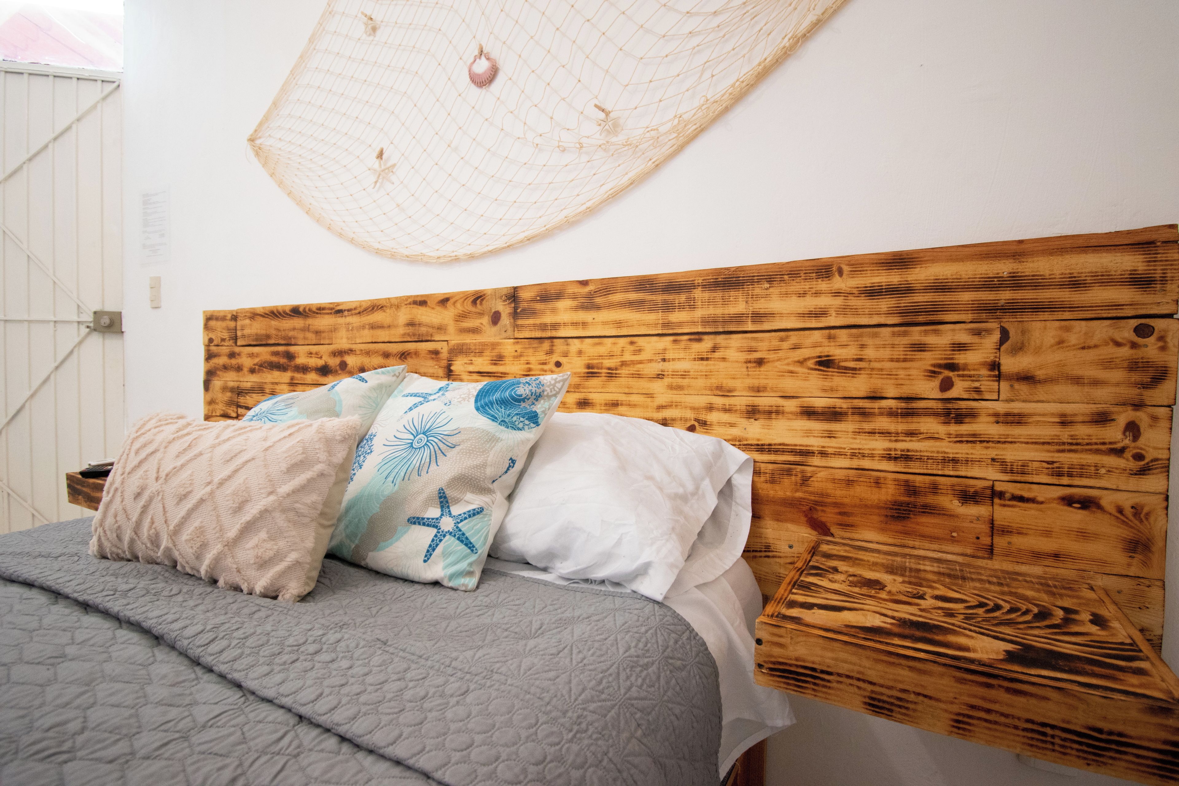 Una cama con colcha gris y varios almohadones, cabecera de madera y mosquitero instalado en la pared.
