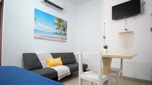 Ett rum med en grå soffa, ett litet vitt bord, en blå säng, en väggmonterad TV och en tavla med ett strandmotiv.