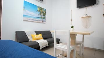 Una habitación con un sofá gris, una mesita blanca pequeña, una cama azul, un televisor montado en la pared y un cuadro con motivos playeros.
