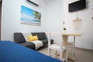 A room with a grey sofa, a small white table, a blue bed, a wall-mounted TV, and a beach-themed painting.