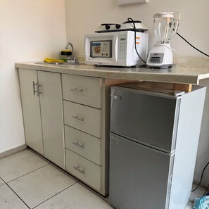 Uma cozinha compacta com micro-ondas, torradeira e liquidificador na bancada, geladeira embaixo e um armário com gavetas.