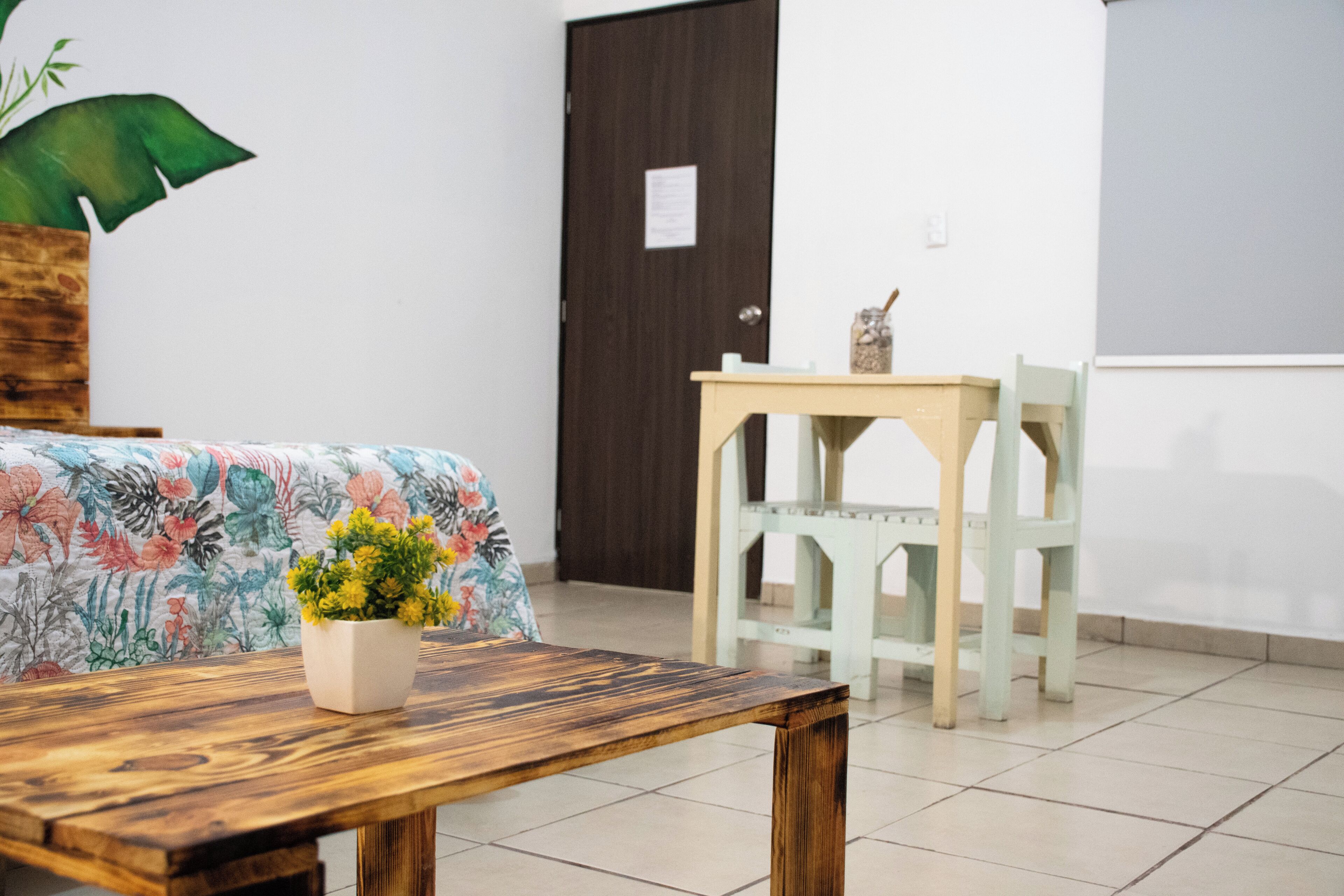 Una habitación con una mesa de madera, una silla, una cama con colcha estampada y una puerta, además de una pared con interruptor de luz.