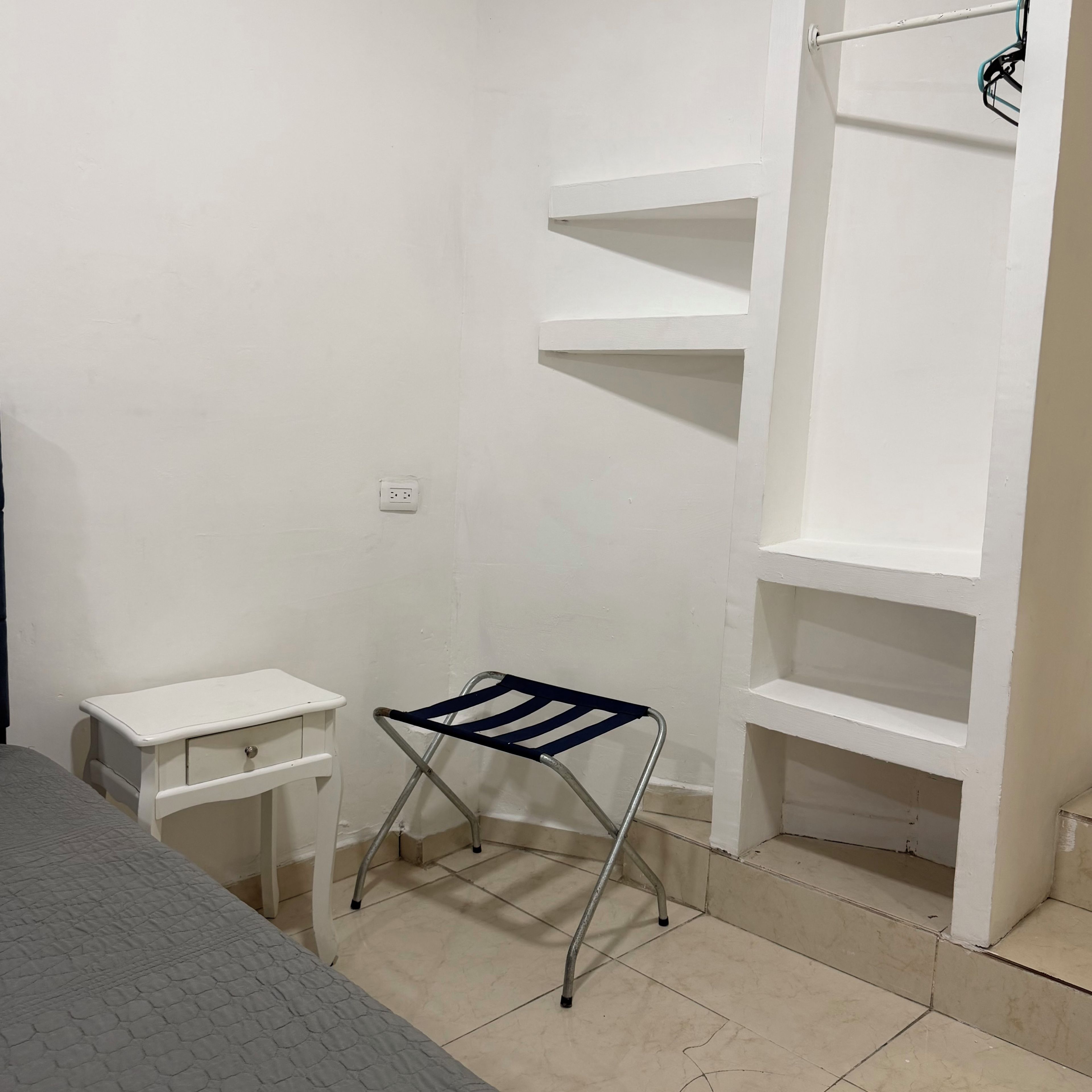 Una habitación con una cama blanca, una mesita de noche pequeña también blanca, una silla plegable de metal, un estante blanco y un suelo de baldosas.