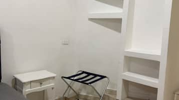Una habitación con una cama blanca, una mesita de noche pequeña también blanca, una silla plegable de metal, un estante blanco y un suelo de baldosas.