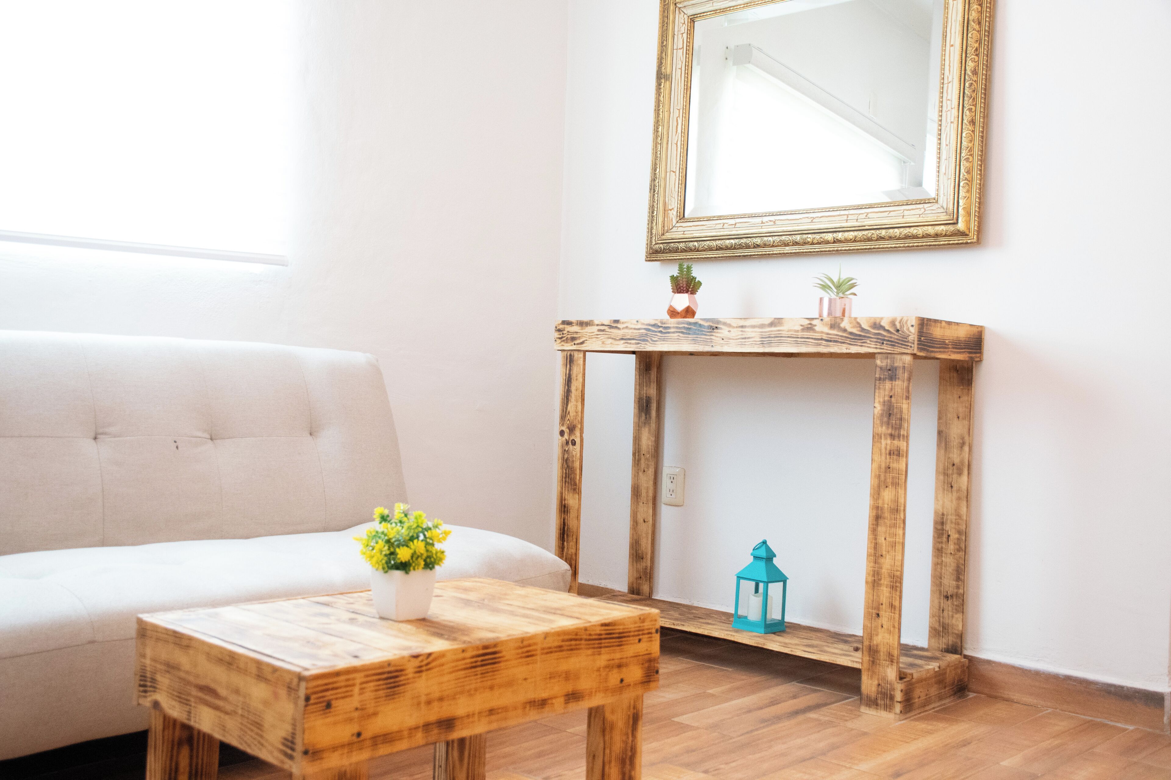 A room with a white sofa, a wooden table with a vase of flowers, a wooden console table, a mirror, and a small plant.